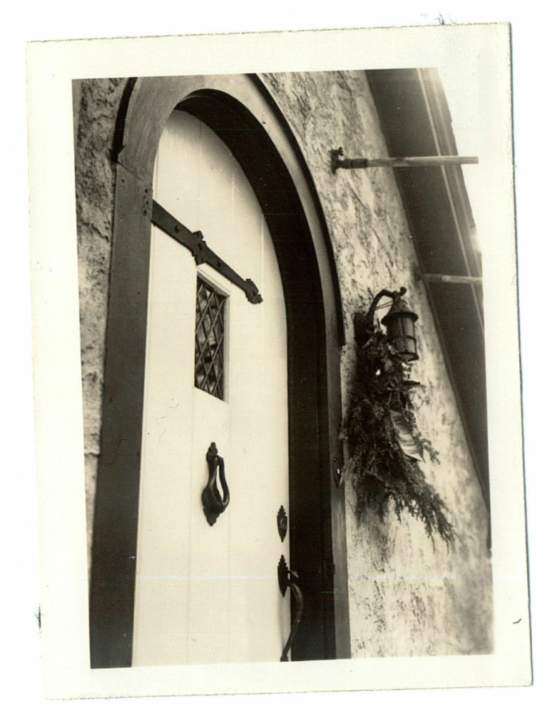 A black and white vintage photo of the white door of a cabin, with diamond-shaped miniature glass panes in the window.