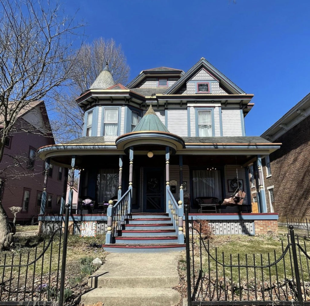 A front view of the Queen Anne Victorian. It has a wrap-around porch with a conical point over the front steps and a wrought-iron fence around the yard.