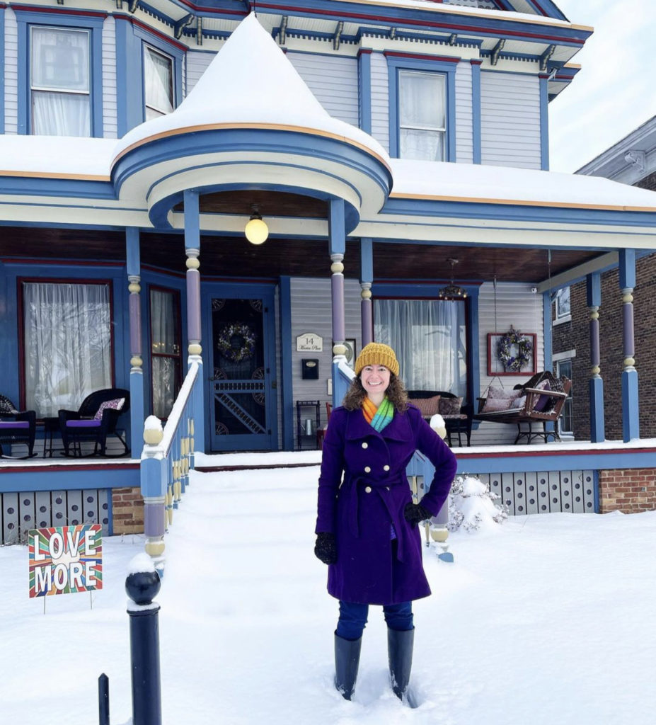 Amy, a light-skinned woman with curly brown hair, posing in a yellow hat and purple coat outside of the Queen Anne in winter. Snow surrounds her and tops the blue and purple porch.