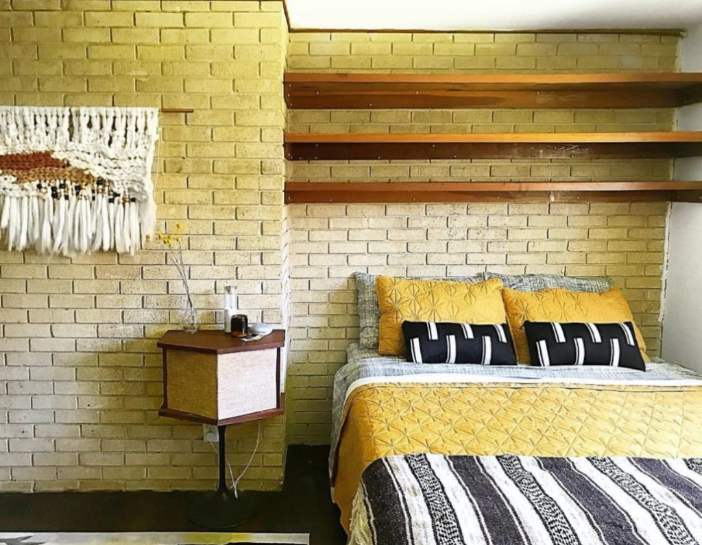 The bedroom of the mid century modern house, with built in shelves over the bed, brick walls, and black and yellow bedding.