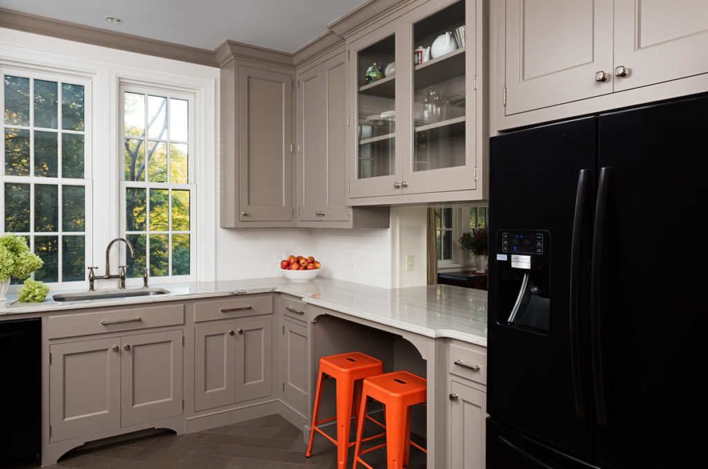 An updated kitchen in an old cottage, with gray trim and white walls and an enormous glossy black fridge. Orange bar stools sit under a breakfast nook.