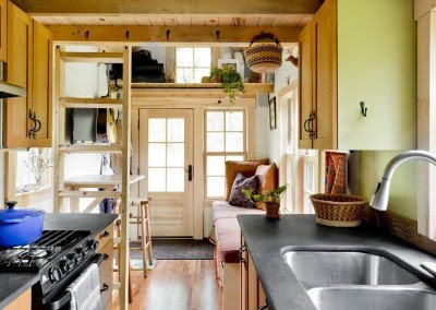 The interior of a tiny house, showing the kitchen and living area. Its windows are bright and most of the surfaces are light-colored wood.