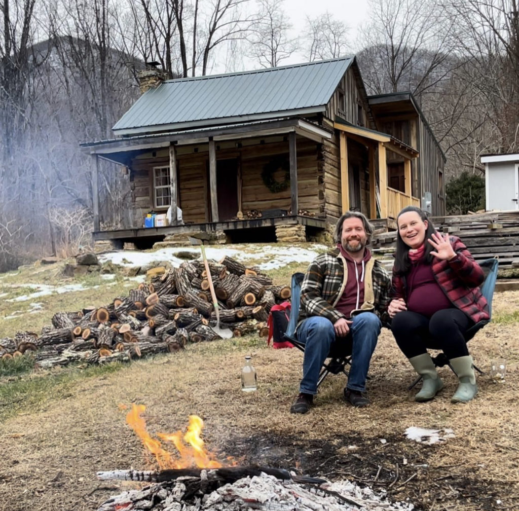 Gabe and Cristina pose in lawn chairs outside their log cabin by a campfire. A pile of firewood sits nearby. They are dressed for warm weather and smiling at the camera.