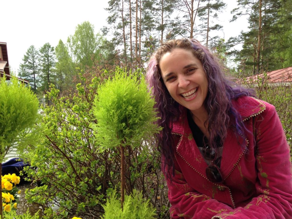 A photo of Tigrilla Gardenia, a woman with tan skin and purple wavy hair. She is wearing a bright pink jacket and posing next to a fluffy bush.