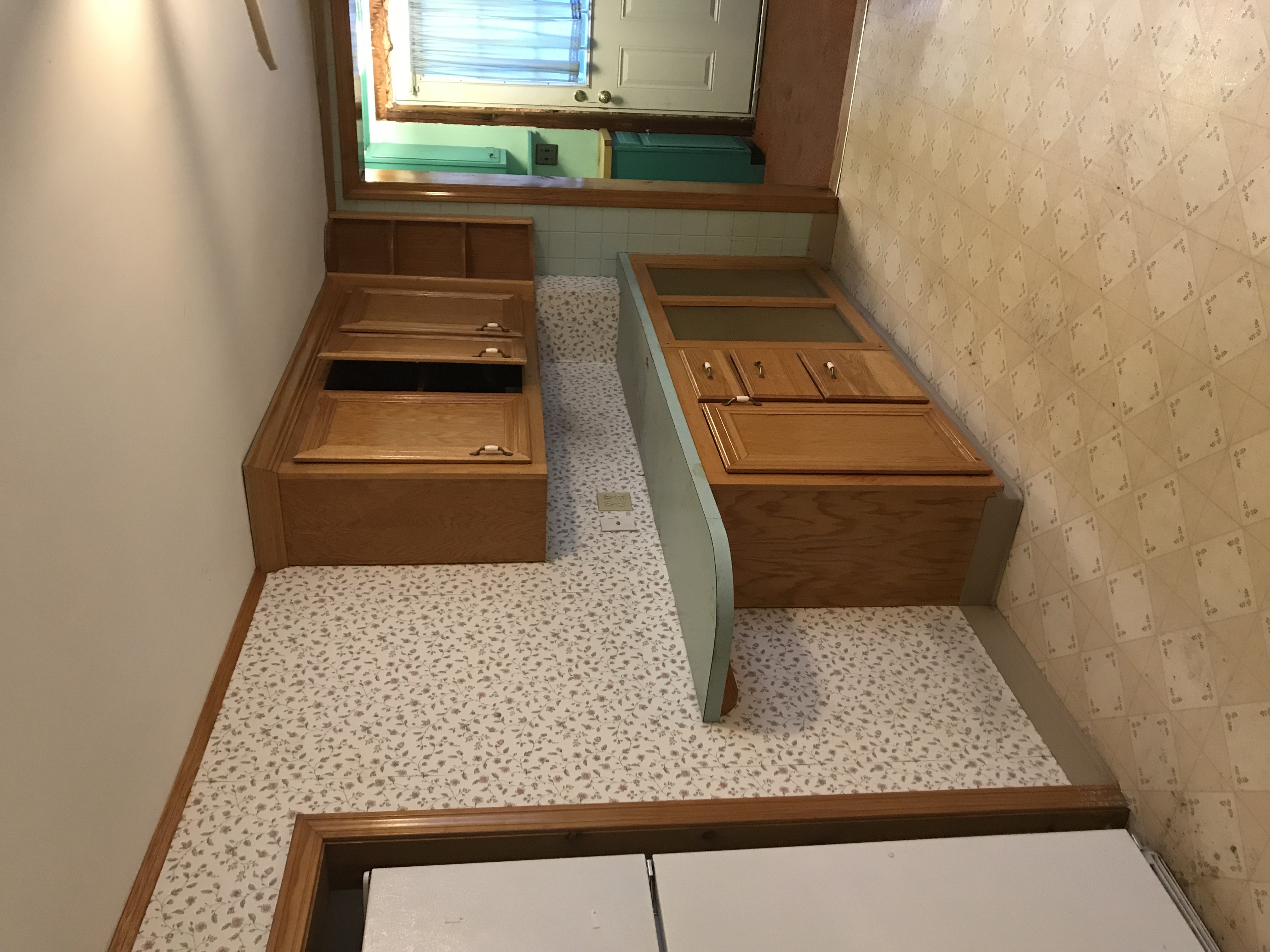 A kitchen during a renovation, with outdated wallpaper, wood cabinets, and Formica countertop. 