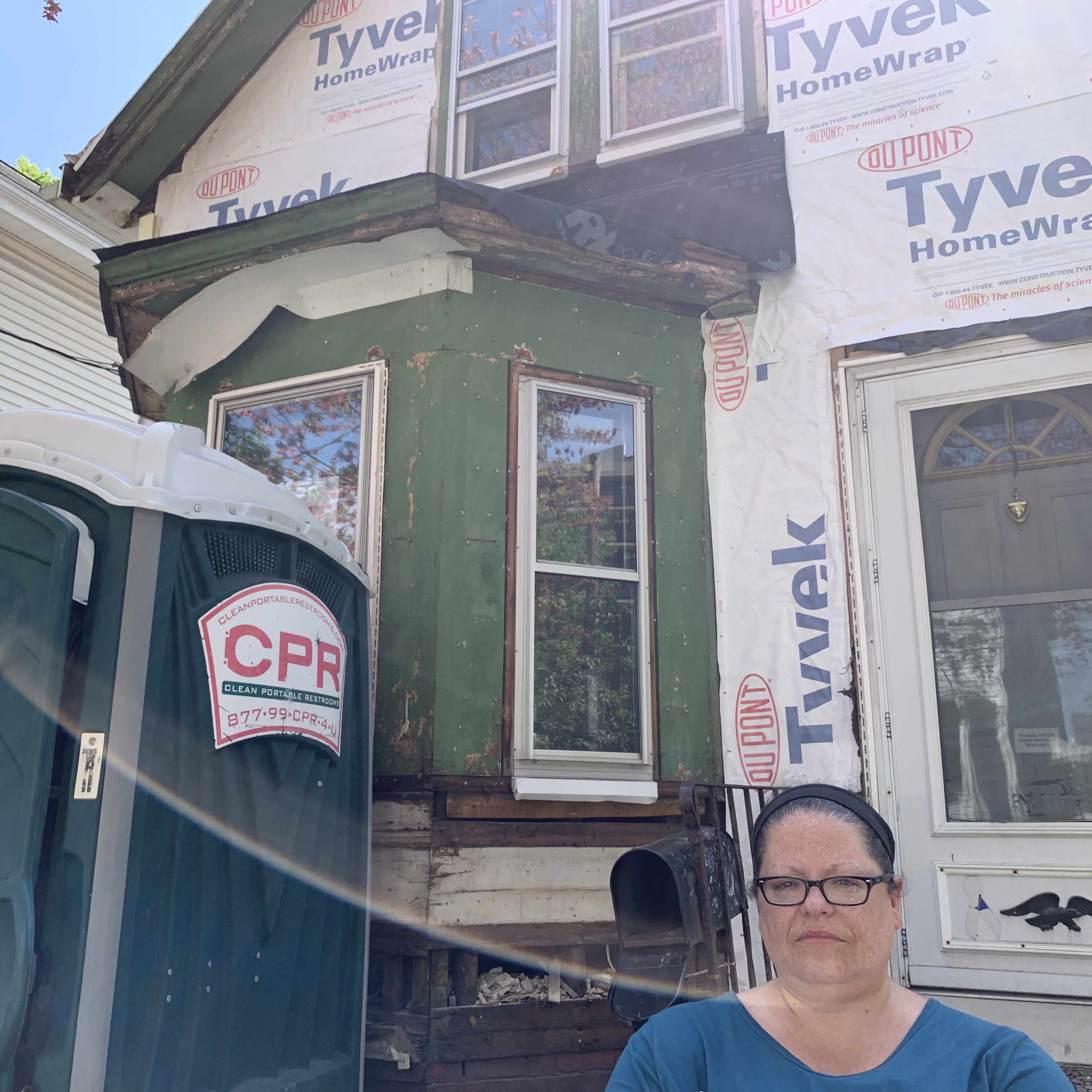 A woman with glasses standing next to her house, which is under construction. There is a portable toilet next to her, and the wall of the house behind her is covered with Tyvek HomeWrap. She is frowning.