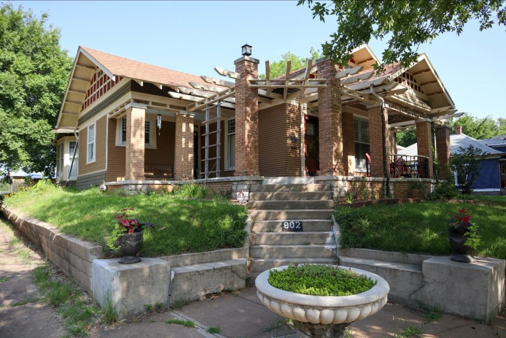 A photo of the front of the Tulsa bungalow after renovation. It has a pergola over the porch and beautiful wood details. There is a fountain out front full of plants.