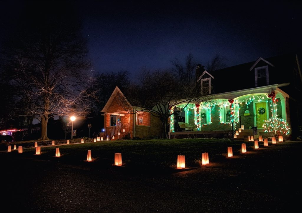 The same old Indianapolis house from the previous photo, this time before the snow, at night. It has paper bag lanterns around the front yard, and the porch is lit up green.