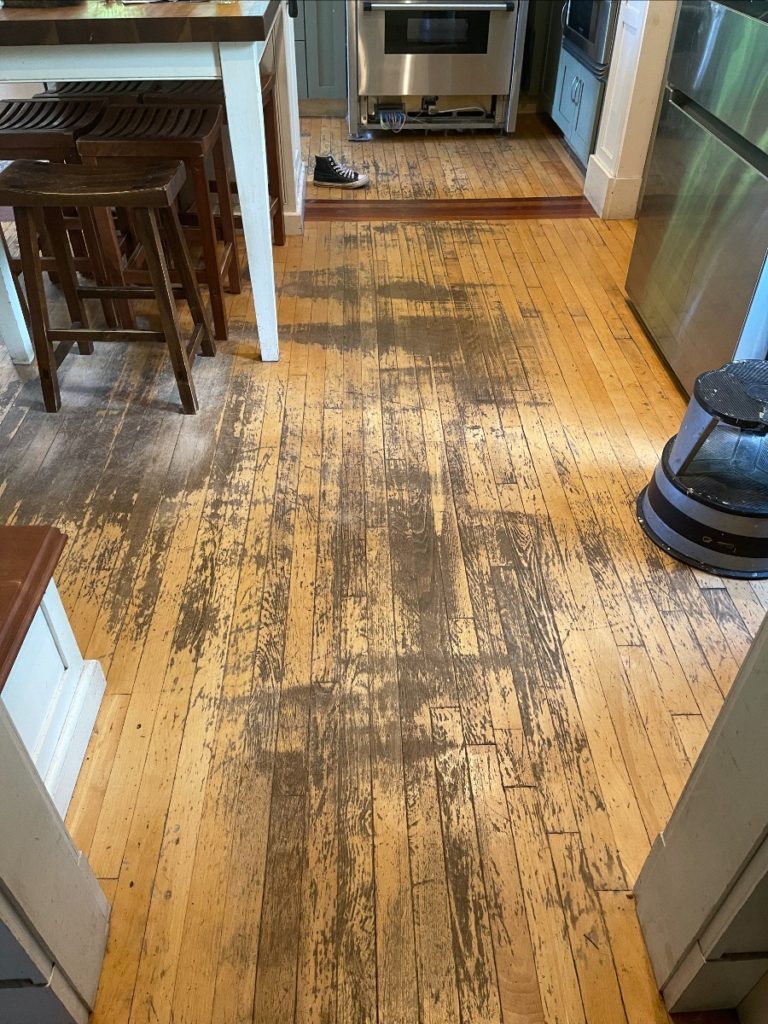 A worn hardwood kitchen floor. The yellowish wood is stained gray and heavily scratched.