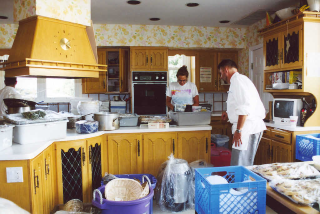 The kitchen before renovation. Two people are standing in the cluttered space. The cabinets are yellowish wood and the walls have orange floral patterned paper.