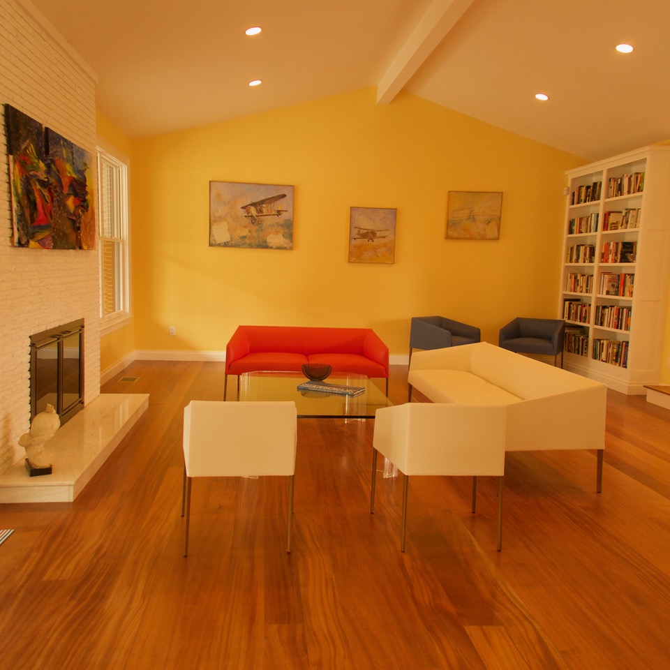 A view of the living room with sleek, geometric-looking red and white couches and a glass coffee table. There is a white fireplace and big piece of abstract art on the left, and a bookshelf that goes almost to the ceiling on the right.