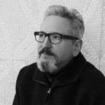 A black and white photo of Mark Ostrom, a light-skinned man with glasses. He has short, graying hair and a graying goatee, and is wearing a dark sweater with a zipper collar.