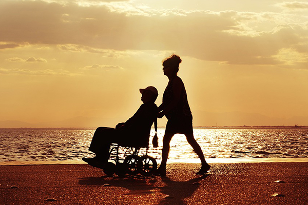 Two people on a beach, silhouetted by the sun. One of the m is pushing the other in a wheelchair.