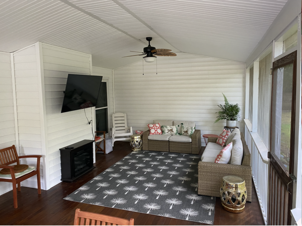 A newly renovated screened porch. There are faux rattan couches, a palm tree patterned rug, and a wall-mounted TV.