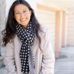 Mona Ying Reeves, a light-skinned woman with long dark hair, smiles at the camera. She is wearing a light-colored sweater and a black and white patterned scarf.