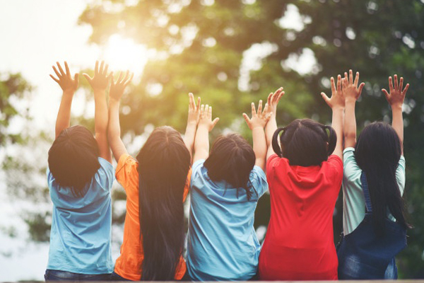 A group of children out in nature. They are facing away from the camera, all reaching into the air together.