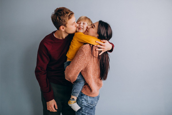 Two parents hugging their young child. One parent is kissing the kid on the cheek.