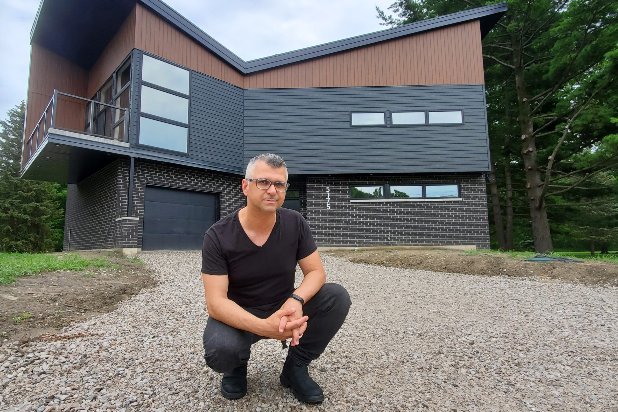 Dory Azar is posing in a squatting position in front of a house he designed, an angular brick structure. He has light skin, gray hair, and glasses, and is wearing black jeans, black shoes, and a black V-neck t-shirt.