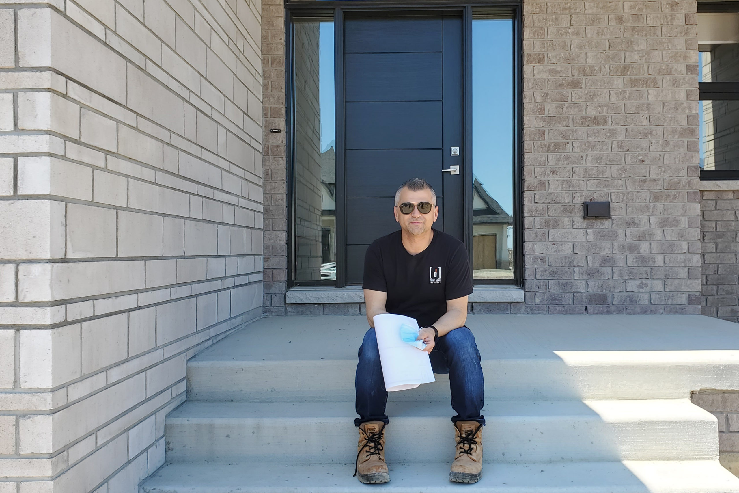 Dory Azar is sitting on concrete stairs. He is wearing sunglasses, jeans, work boots, and a black t-shirt.