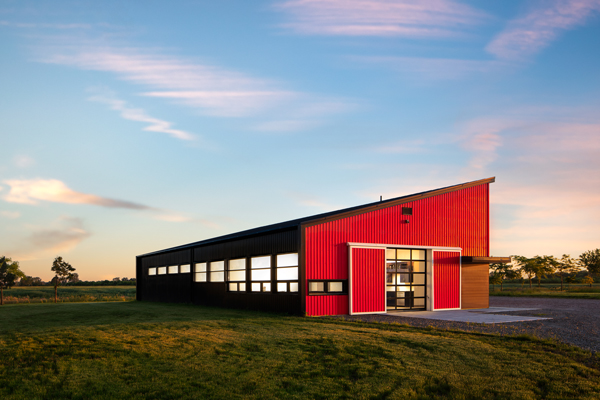 A striking, angular, modern one-story house with red siding, designed by Dory Azar. Golden sunlight hits the side of the building while pink clouds pass through a blue sky overhead.