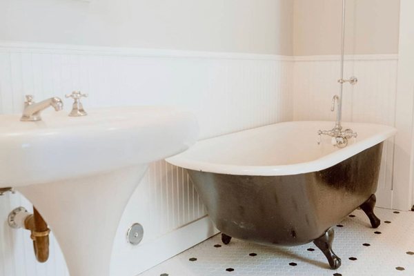 Part of a project to restore a historic home: a classic vintage bathroom: clawfoot bathtub with antique fixtures, an old-fashioned sink, and a white hexagonal tile floor with black accent tiles. The walls are white beadboard.