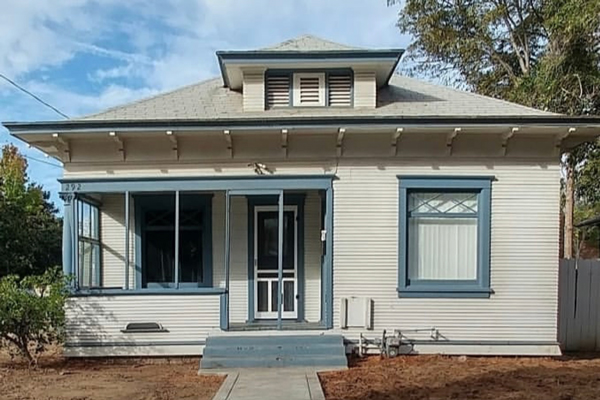 A one-story historic home with white paint and blue trim. There is a small porch. The yard is empty dirt.