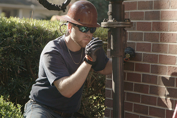 A foundation professional wearing a hard hat and gloves. He is examining a large jack outside of a house with a brick exterior.