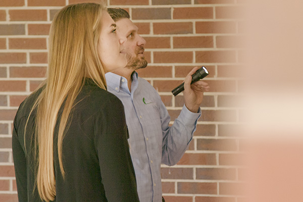 Two light-skinned people, a man and a woman, examining an exterior wall for cracks that would be evidence of foundation problems. The man is holding a flashlight. 