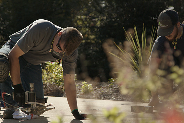 Two repair professionals inject foam beneath a sidewalk to stabilize the soil and keep the concrete level.