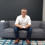 Dory Azar, a light-skinned man with glasses, is posing in business casual dress on a small gray couch.