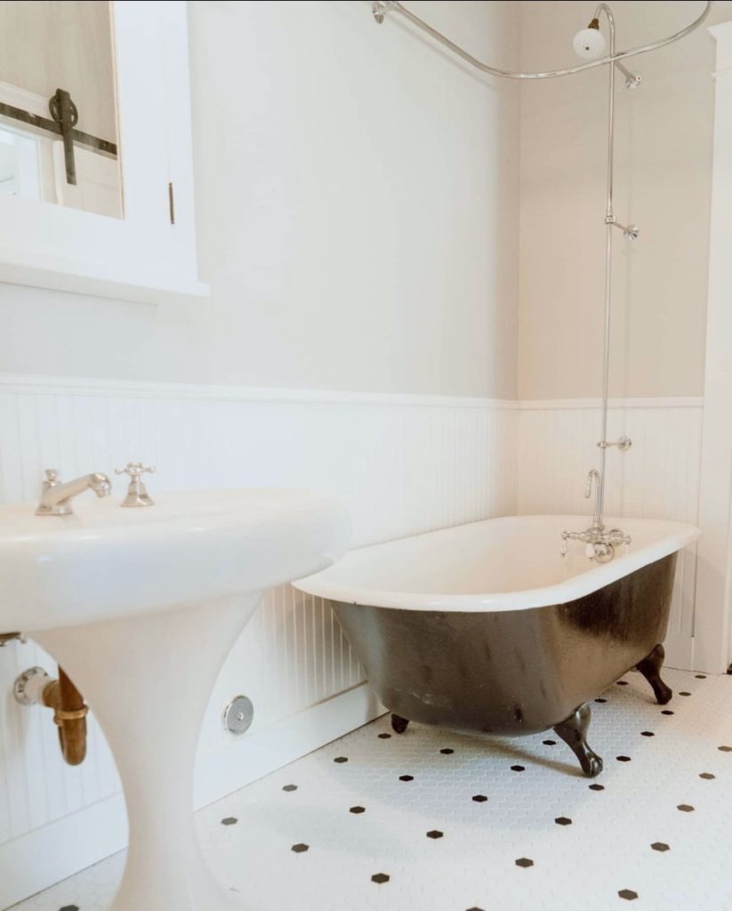 Inside a historic home restoration, a classic vintage bathroom: clawfoot bathtub with antique fixtures, an old-fashioned sink, and a white hexagonal tile floor with black accent tiles. The walls are white beadboard.