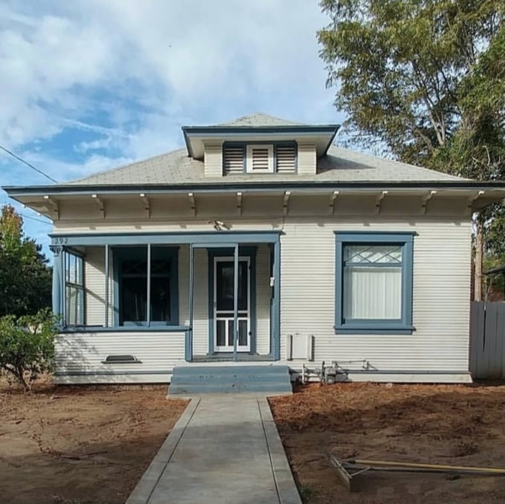 A one-story historic home with white paint and blue trim. There is a small porch. The yard is empty dirt.