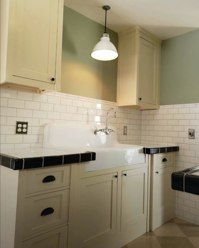 A historic kitchen restoration. Black and white subway tile, an antique sink and vintage light fixture, and sage green walls above checkered floor tiles.