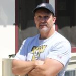 A photo of Hugh Siler. He is a muscular, light-skinned man wearing a t-shirt and a baseball cap. He is squinting as he smiles at the camera.